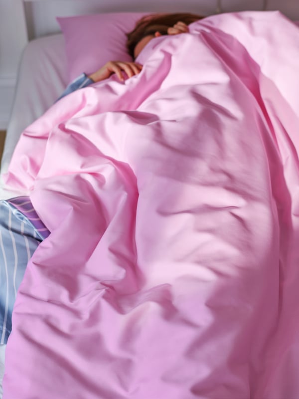 Close-up of person in pajamas sleeping under a bright pink sheet that is pulled up over the face. Picture taken from above.