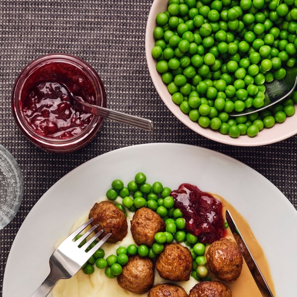 A white plate has a portion of meatballs, mashed potatoes and green peas, with a jar of lingonberries next to it.