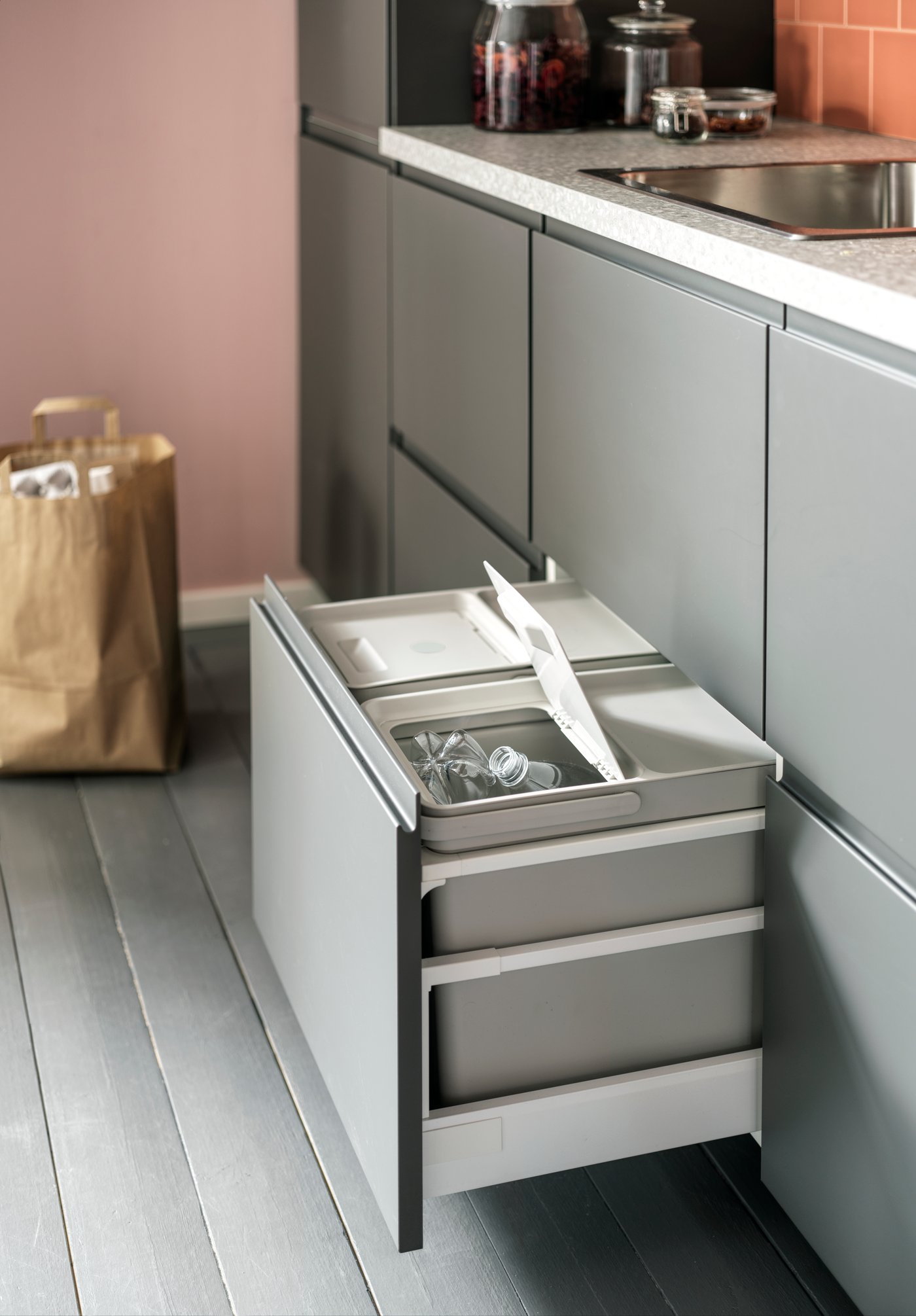An open kitchen drawer with light grey waste sorting bins inside. One lid is open and plastic bottles are sorted inside.