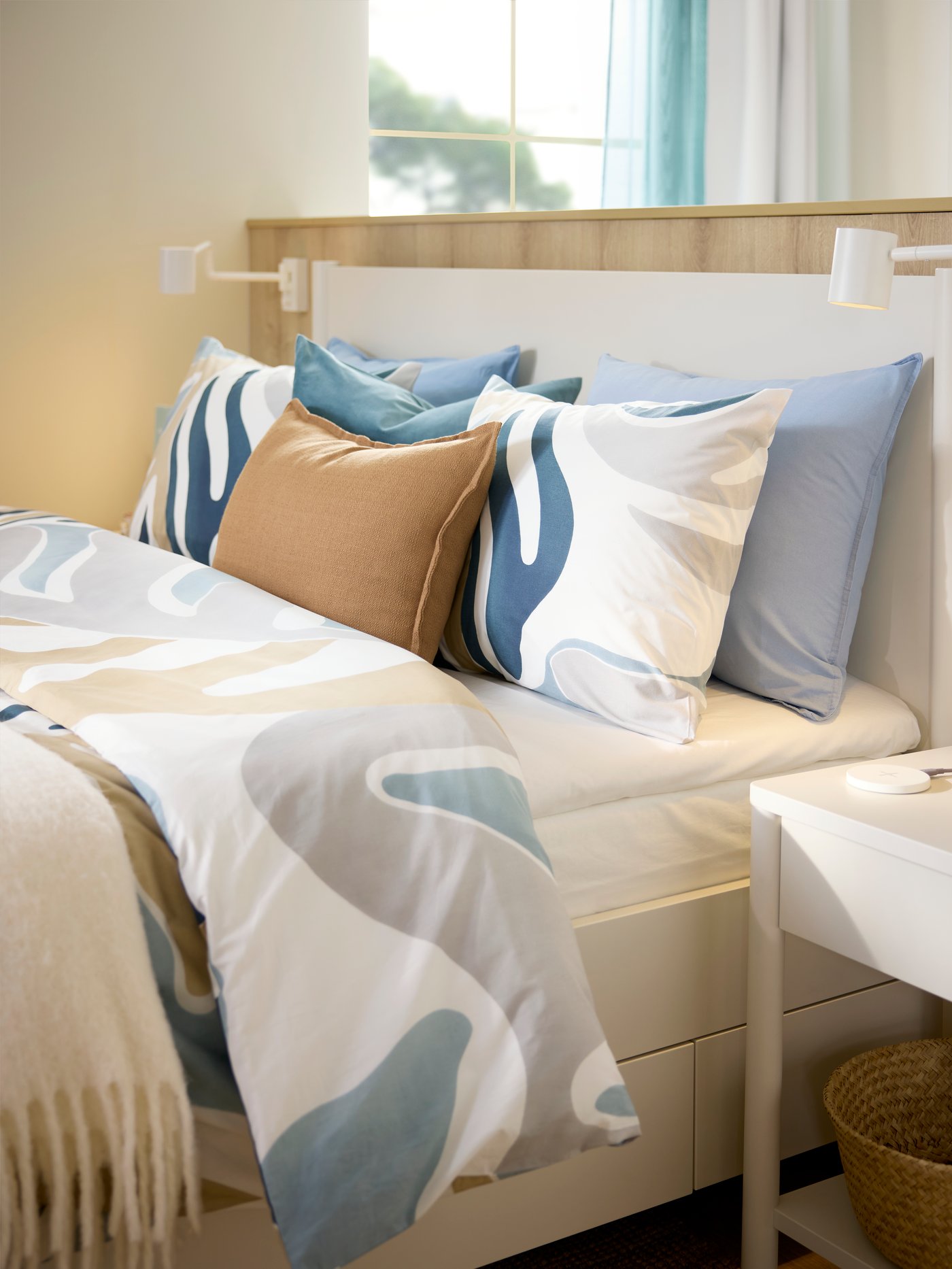 A hotel bed by a low wooden dividing wall is made with MYGGLASVINGE duvet cover and pillowcases that has a big leafy pattern.