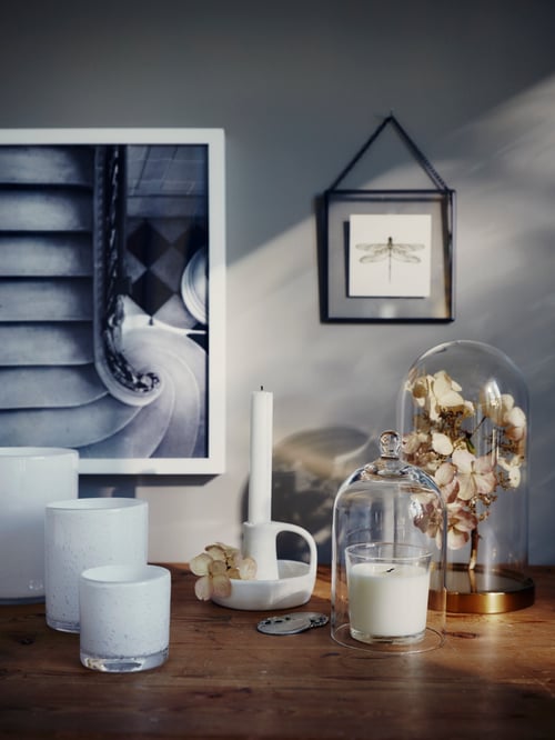 Tabletop with a glass dome with base, multiple candle holders, and a candlestick, and a poster and frame on the wall.