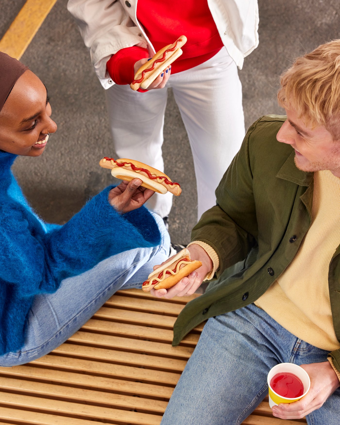To personer sidder på en bænk de holder hotdogs i hænderne og den ene et glas med saft.