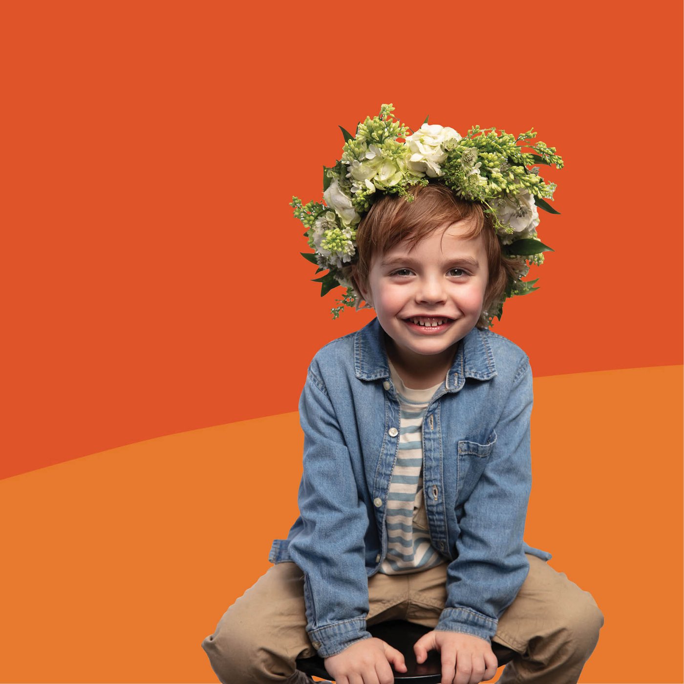 Enfant avec couronne de fleurs assis sur un tabouret sur fond orange, tenue décontractée et ambiance ludique