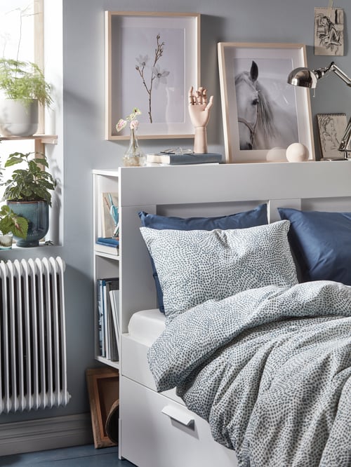 A bed made with white-and-blue TRÄDKRASSULA bed linen. Framed art above the bed, and plants in a nearby window.