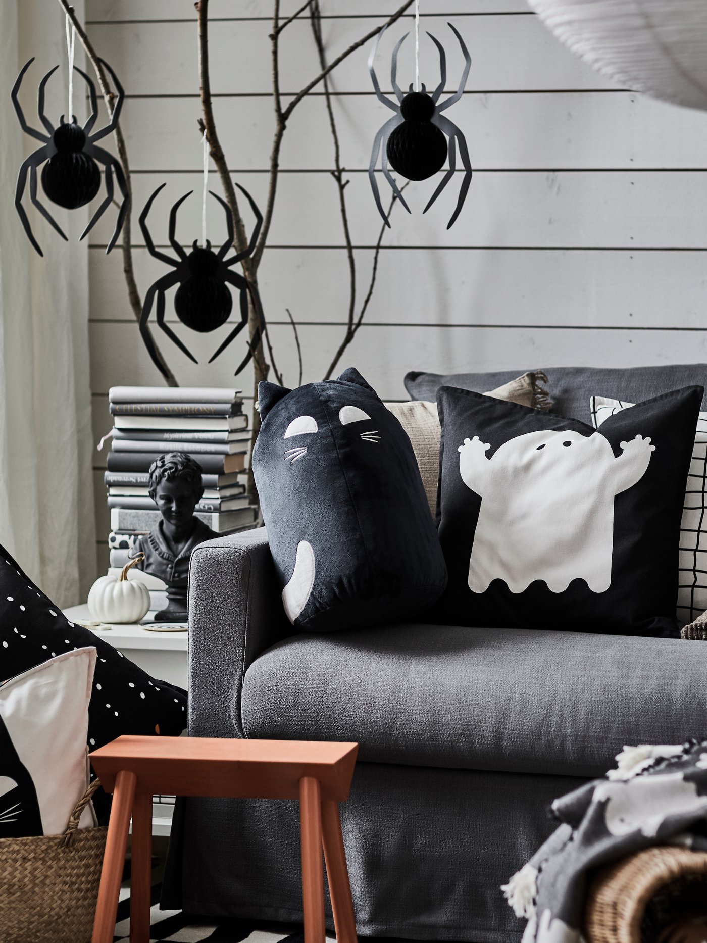 A living room with a grey sofa, KUSTFYR cushion covers featuring ghosts and black cats, and hanging decorations of spiders.