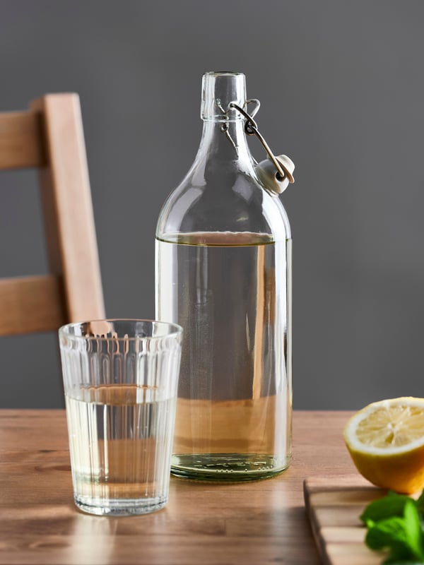 A KORKEN bottle with stopper in clear glass is placed on a wood table next to a glass and a cutting board with half a lemon.