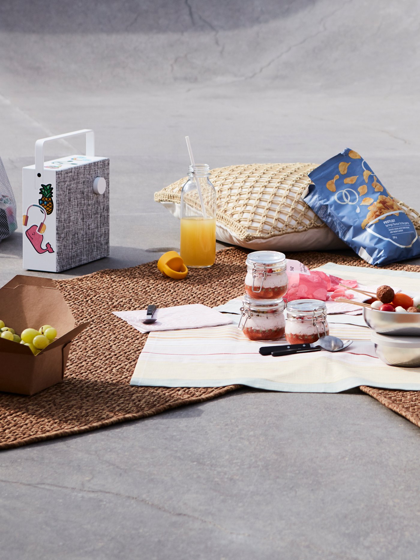 Décor de pique-nique dans un cadre urbain avec desserts dans des bocaux en verre, jus, chips, tapis d’extérieur, enceinte et coussins. 