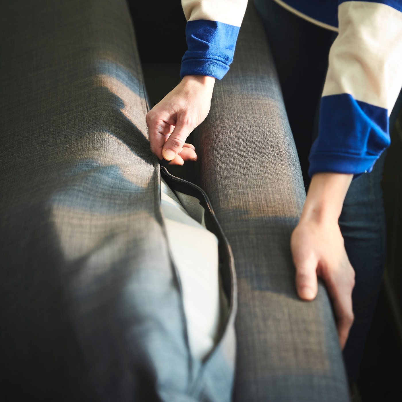 A person pulls the zipper of a dark-grey KLAGSHAMN back-cushion cover of a dark-grey FRIHETEN corner sofa-bed.