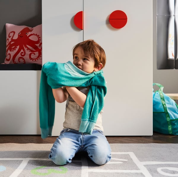 A child getting dressed on a grey rug in front of a white bench with toy storage and a white wardrobe with a pull-out unit.