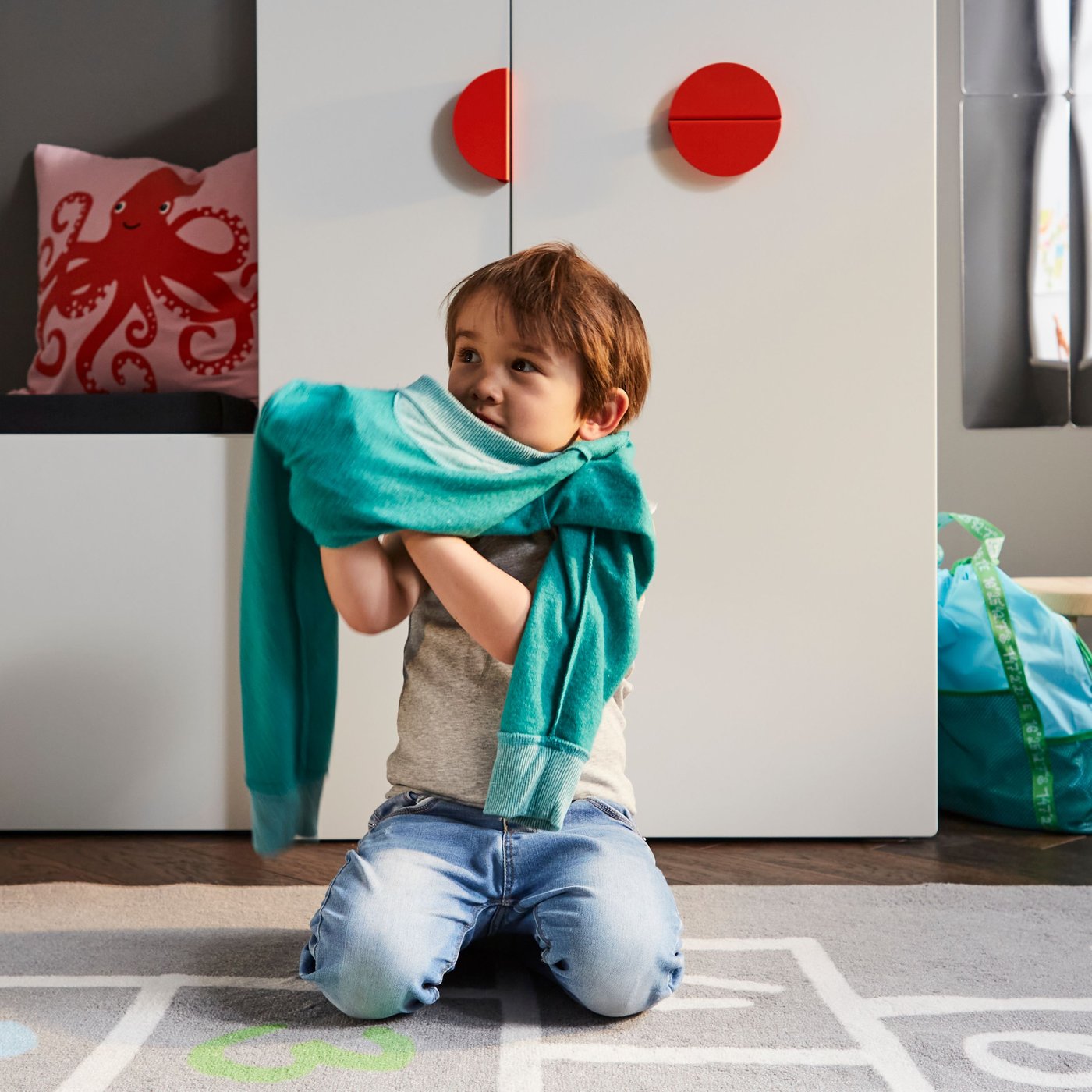 A child getting dressed on a grey rug in front of a white bench with toy storage and a white wardrobe with a pull-out unit.