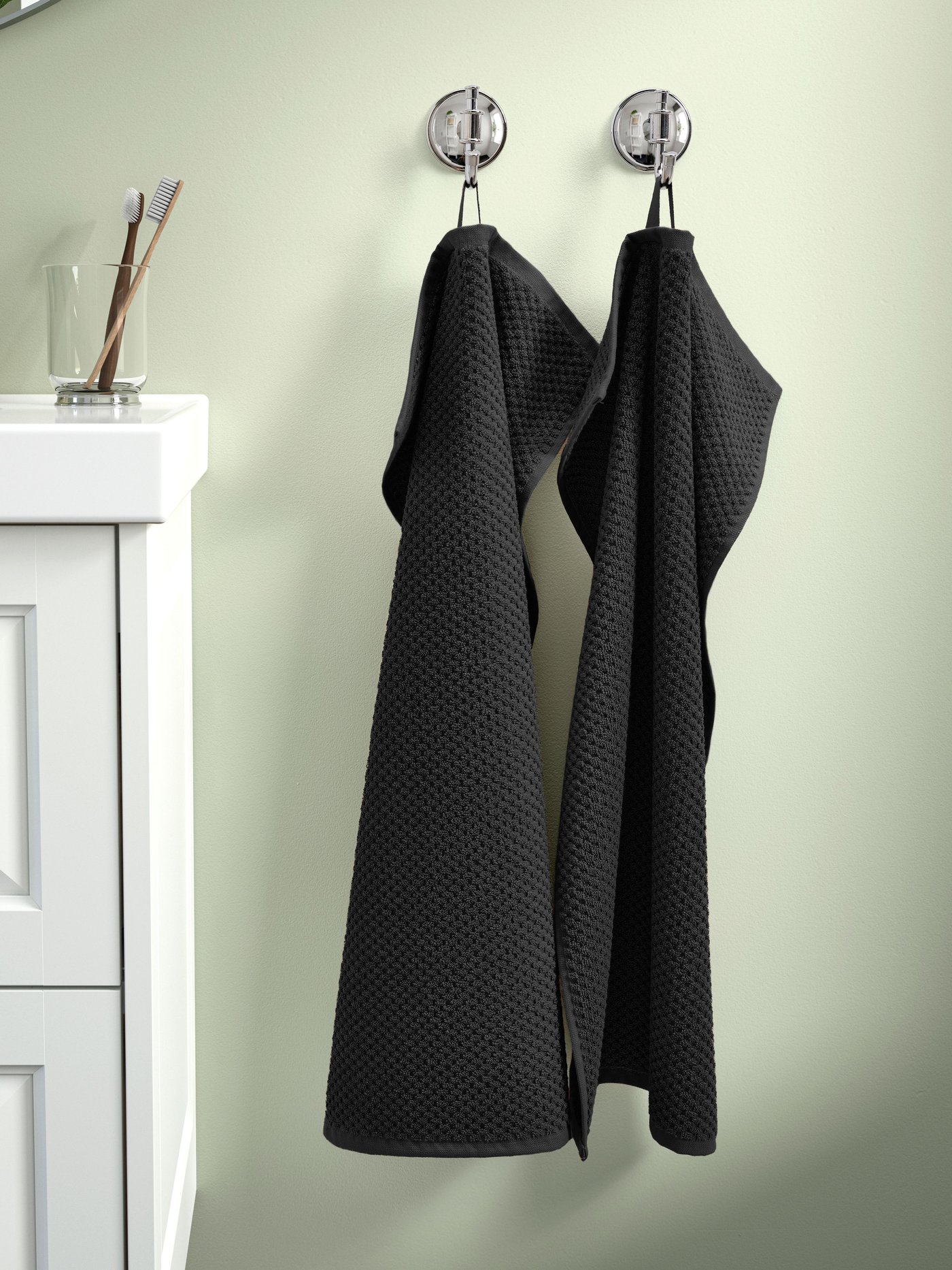 Czarne ręczniki GULVIAL wiszą na srebrnych haczykach na zielonej ścianie.