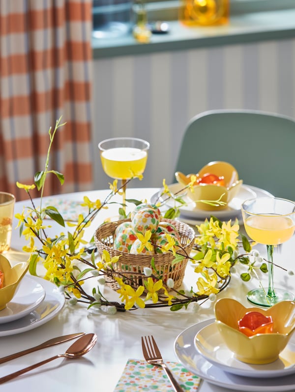 Bright spring table setting with a basket filled with coloured eggs and a yellow flowered wreath around it.