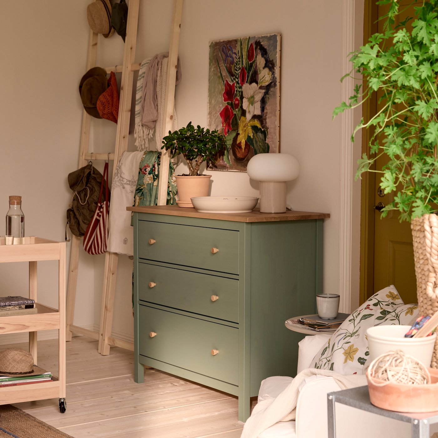 En grøn HEMNES kommode med potter og planter ovenpå, et billede over og træstiger bagi til opbevaring.