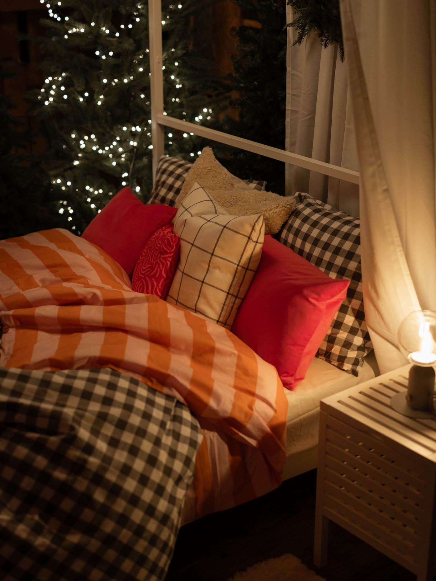Säng med orange- och vitrandigt täcke, kuddar i olika färger och mönster. I bakgrunden en julgran med ljusslingor.

