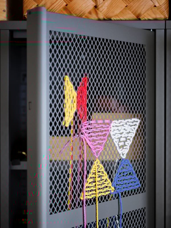 A section of a grey IVAR cabinet where colourful threads have been used to embroider decorations on the door's metal mesh.