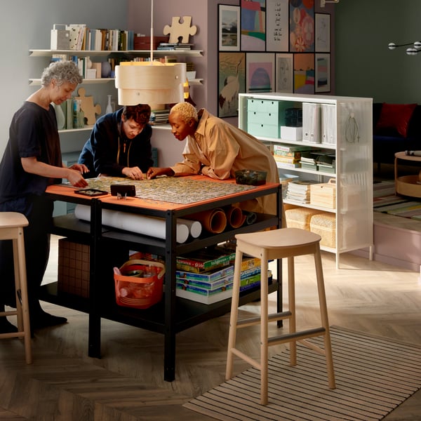 Three people by table looking at fabric under wooden lamp in well-organised home full of colours.