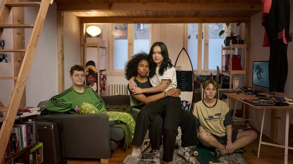 Four people sits in a room filled with furnishings and look straight into the camera.