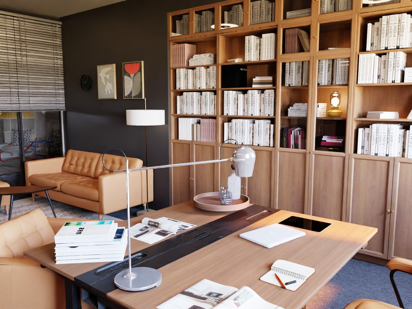 Teil eines Büros. Blick auf eine Wand mit Bücherregalen. Davor ein Schreibtisch mit zwei Stühlen. Auf dem Schreibtisch Dokumente und eine Leseleuchte. Im Hintergrund rechts ein Zweisitzersofa und davor ein Beistelltisch. An der Wand über dem Sofa ein Bild und neben dem Sofa eine Standleuchte.