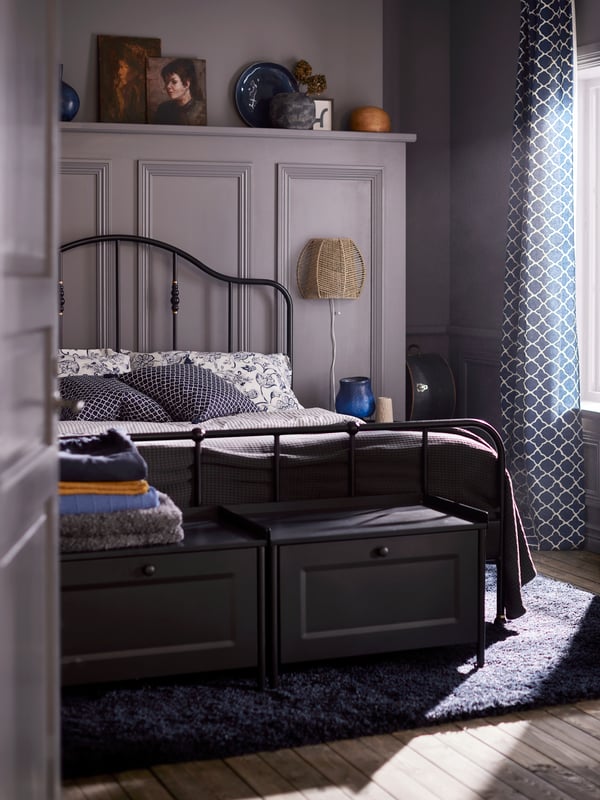 Two black storage benches and a bed with a black SAGSTUA bed frame made with white/blue floral bed linen on a dark blue rug.