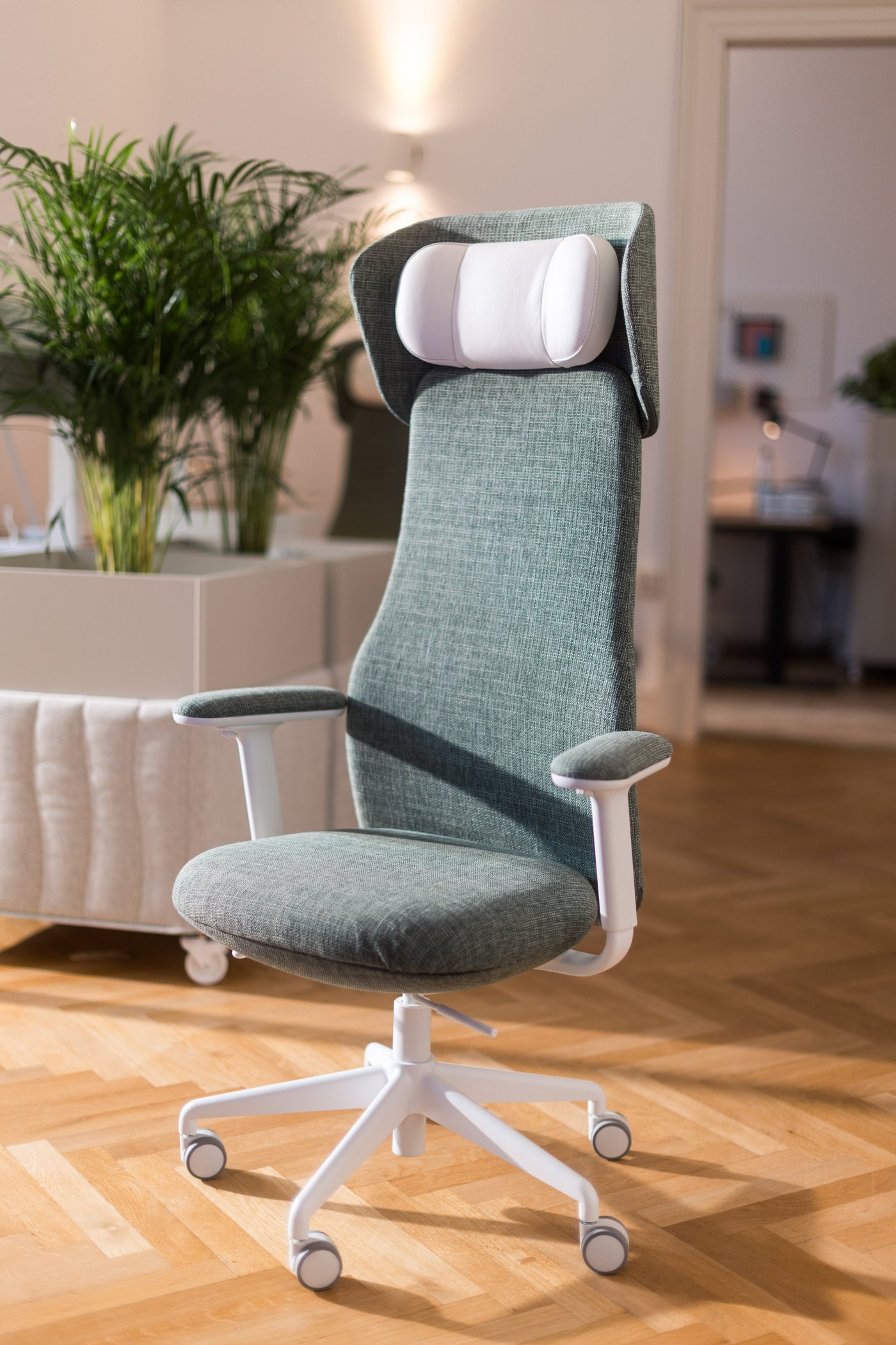 Ergonomischer Bürostuhl mit hoher Lehne und Nackenkissen, vor Pflanzen und Arbeitsplätzen im Hintergrund.