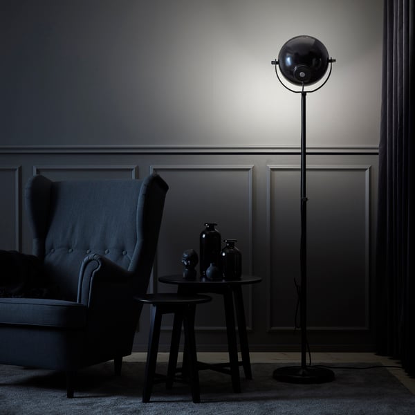 Mörkt, stämningsfullt vardagsrum med en hög svart golvlampa som lyser mot en grå panelvägg, bredvid en mörk fåtölj och två små runda bord med svarta dekorativa vaser.