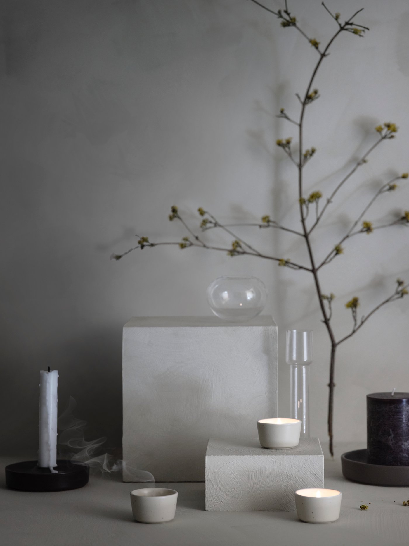 Trois photophores ÄROFULL beiges sont présents dans un agencement minimaliste de nature morte avec des verres, des bougies et du feuillage.