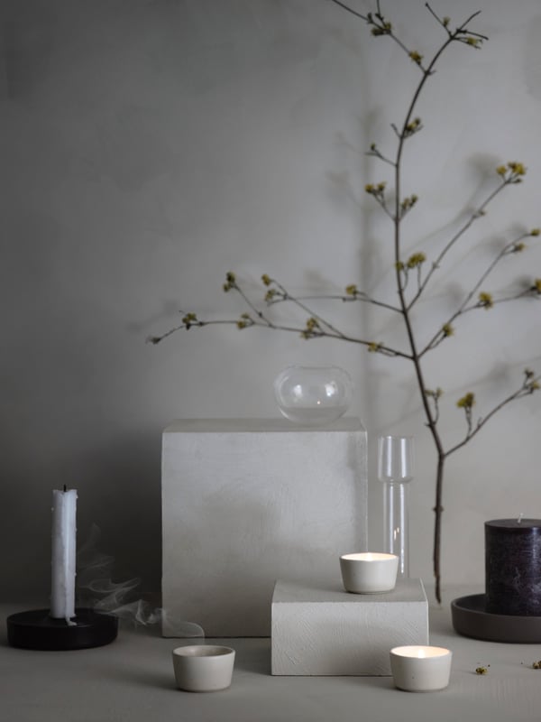 Three beige ÄROFULL tealight holders are displayed in a minimal still life setting with glassware, candles and foliage.