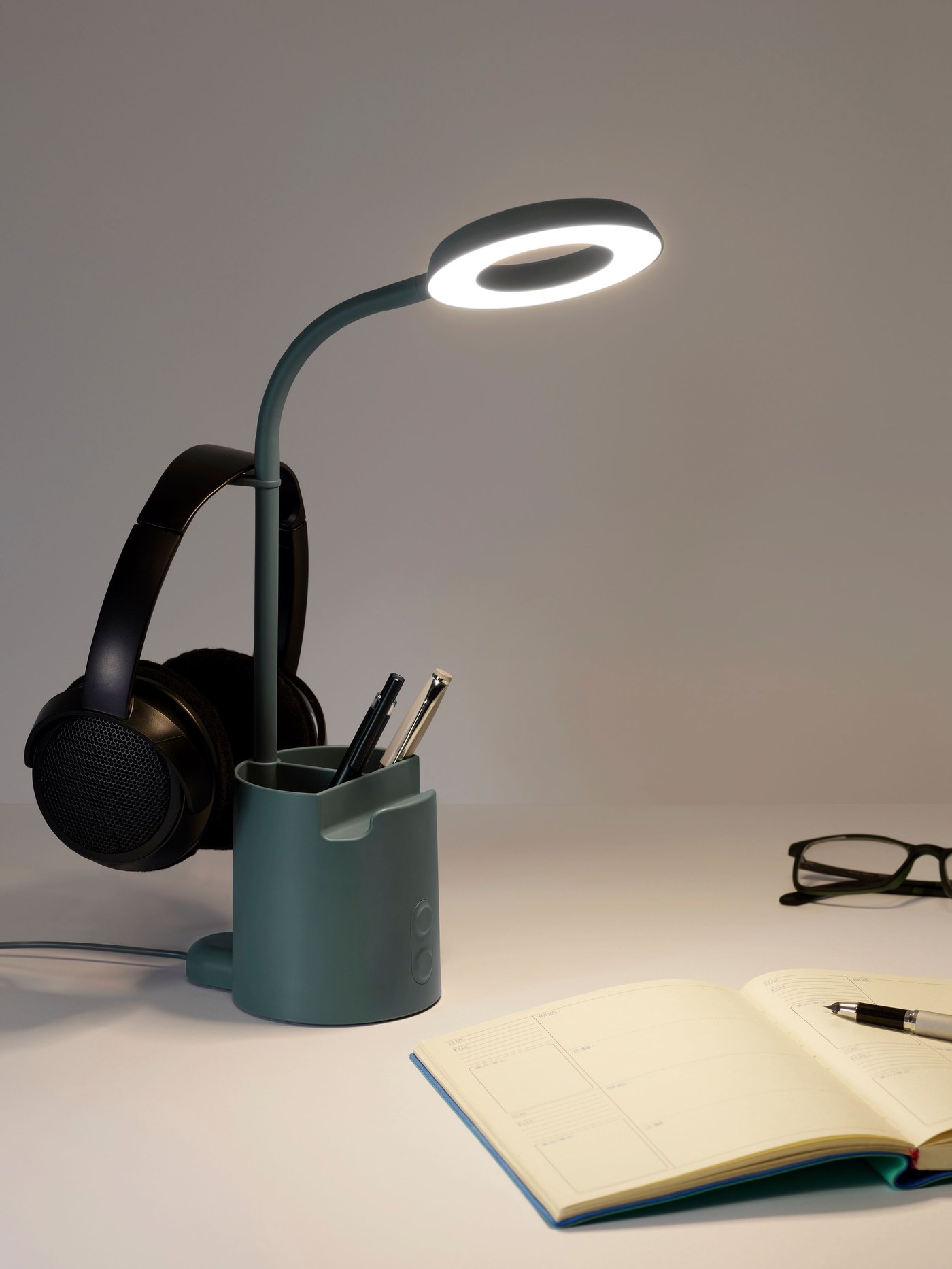 BRUNBÅGE LED-arbetslampa med förvaring, dimbar/turkos. Det hänger ett par hörlurar på lampan och på skrivbordet ligger ett öppet anteckningsblock och ett par glasögon.