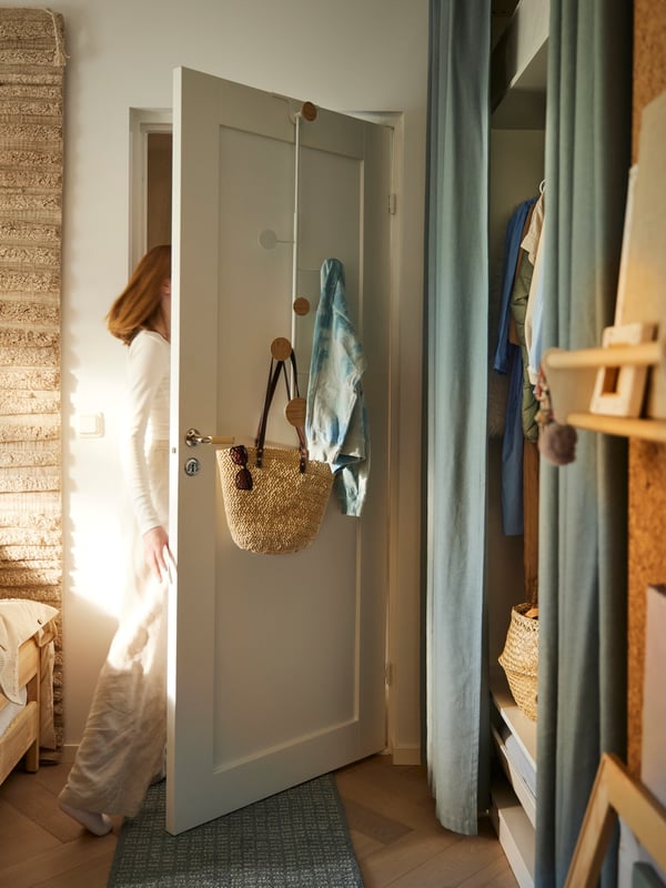 Pessoa a sair de quarto por porta em branco com cabide para porta BÄRFIS com saco e roupa pendurados.