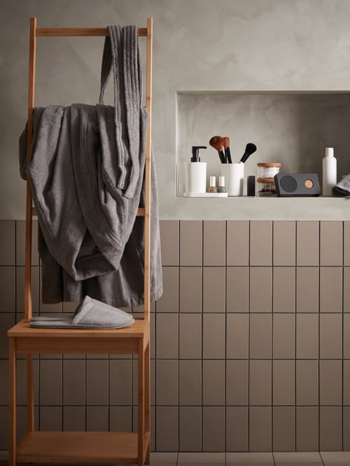 Une chaise en bois avec des chaussons et un peignoir gris se trouve à côté d’une étagère avec un ensemble d’accessoires de salle de bain STORAVAN et une enceinte ENEBY.