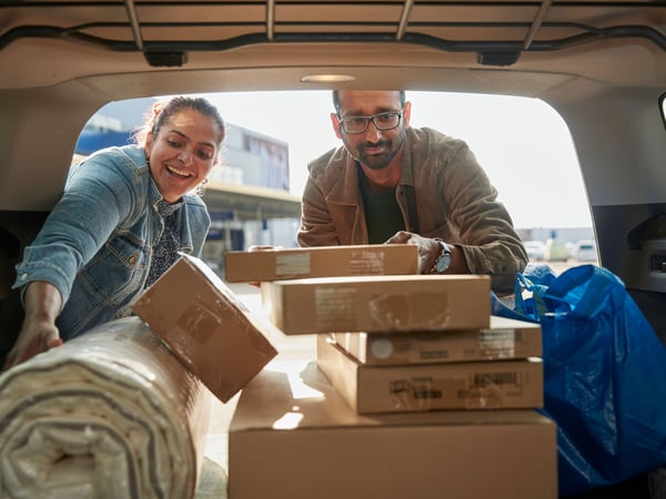 Deux personnes chargent et rangent des cartons de meubles en kit et un tapis enroulé dans le coffre d'une voiture après récupération d'articles d'ameublement achetés.