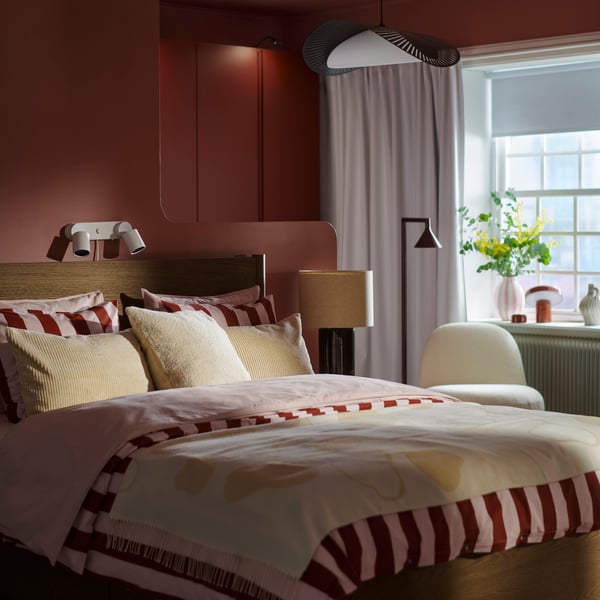 Ein Schlafzimmer mit einem dunkelbraunen TONSTAD-Bett, bezogen mit gestreifter SLÅNHÖSTMAL-Bettwäsche sowie cremefarbenen Kissen und einer Tagesdecke, im Hintergrund ein Fenster. An der Decke über dem Bett hängt eine KVÄLLSDOPP Leuchte.