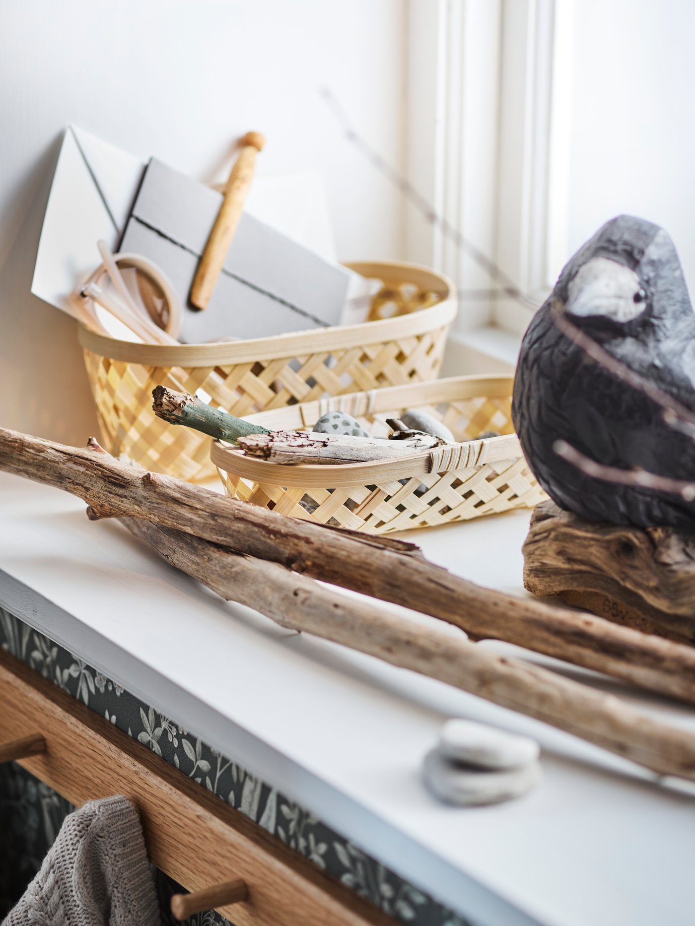 A set of two SALUDING baskets, stones, sticks and a bird ornament are on a windowsill above a HÖVOLM rack with 6 knobs.