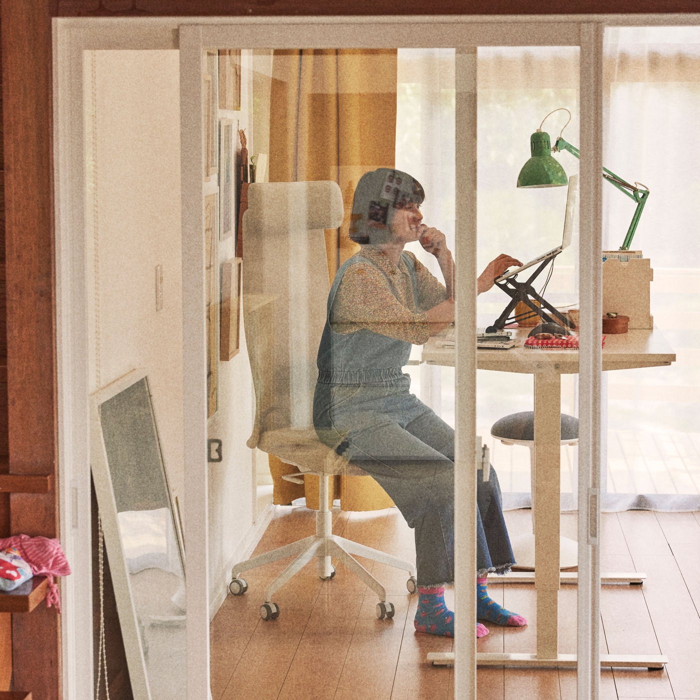 A woman sits on an office chair at a desk with a window in front.
