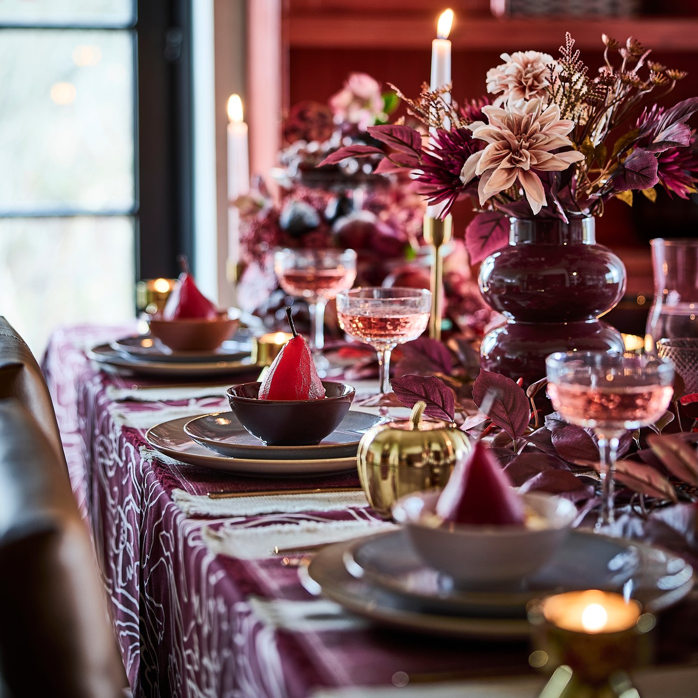 Una mesa con decoración rosa y roja, velas encendidas, ramos de flores artificiales HÖSTAGILLE en jarrones y peras escalfadas en cuencos.