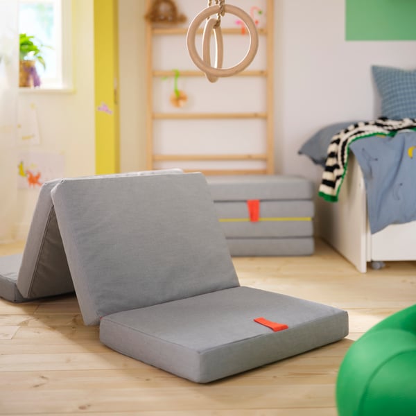 An unfolded SLÄKT pouffe/mattress is placed on the floor in a room with wall bars and gymnastic flying rings.
