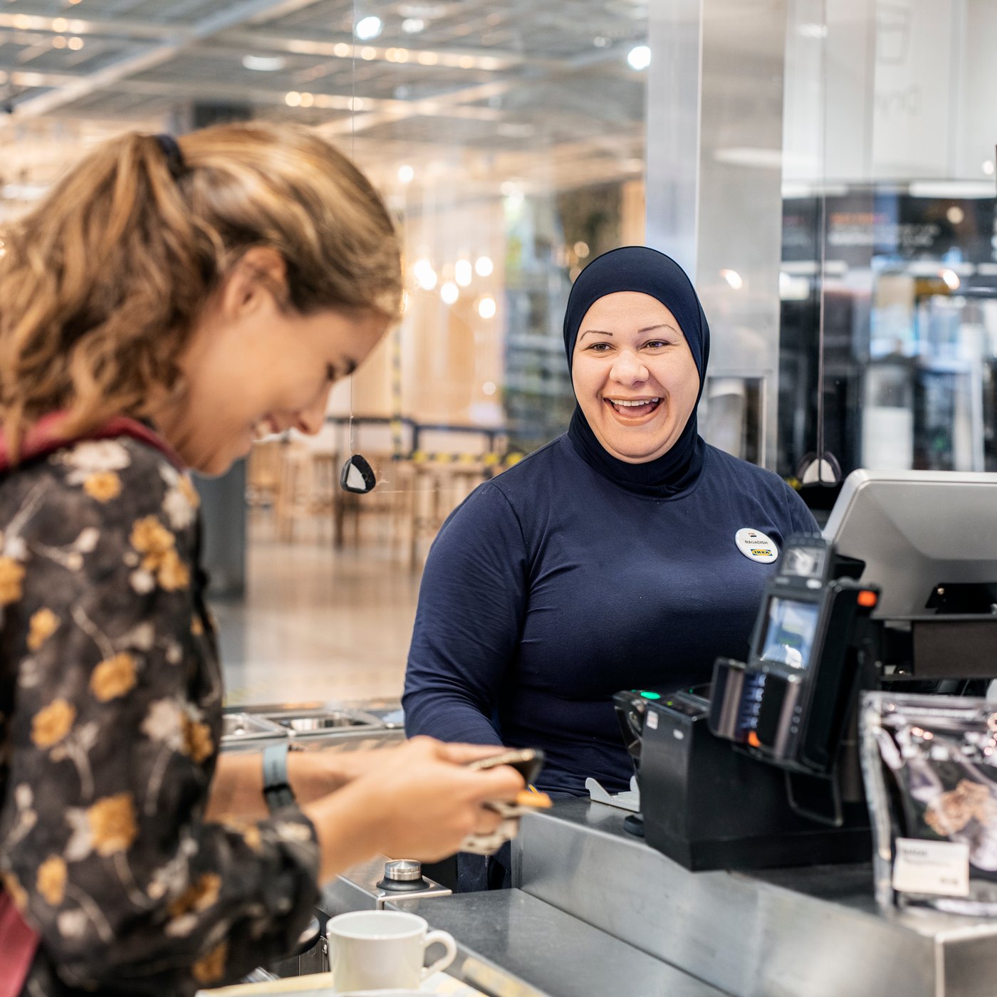 Hijabia käyttävä kassatyöntekijä nauraa asiakkaan kanssa kassan äärellä.
