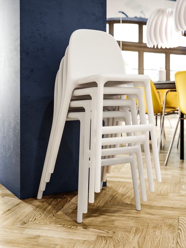 Des chaises d’enfants blanches empilées contre un mur bleu, un plancher en bois, des suspensions blanches et des chaises jaune foncé en arrière-plan.