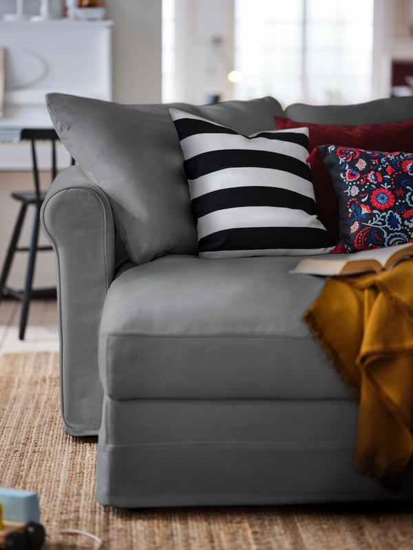 A living room where various colourful cushions and a throw occupy the chaise longue part of a grey GRÖNLID four-seat sofa.