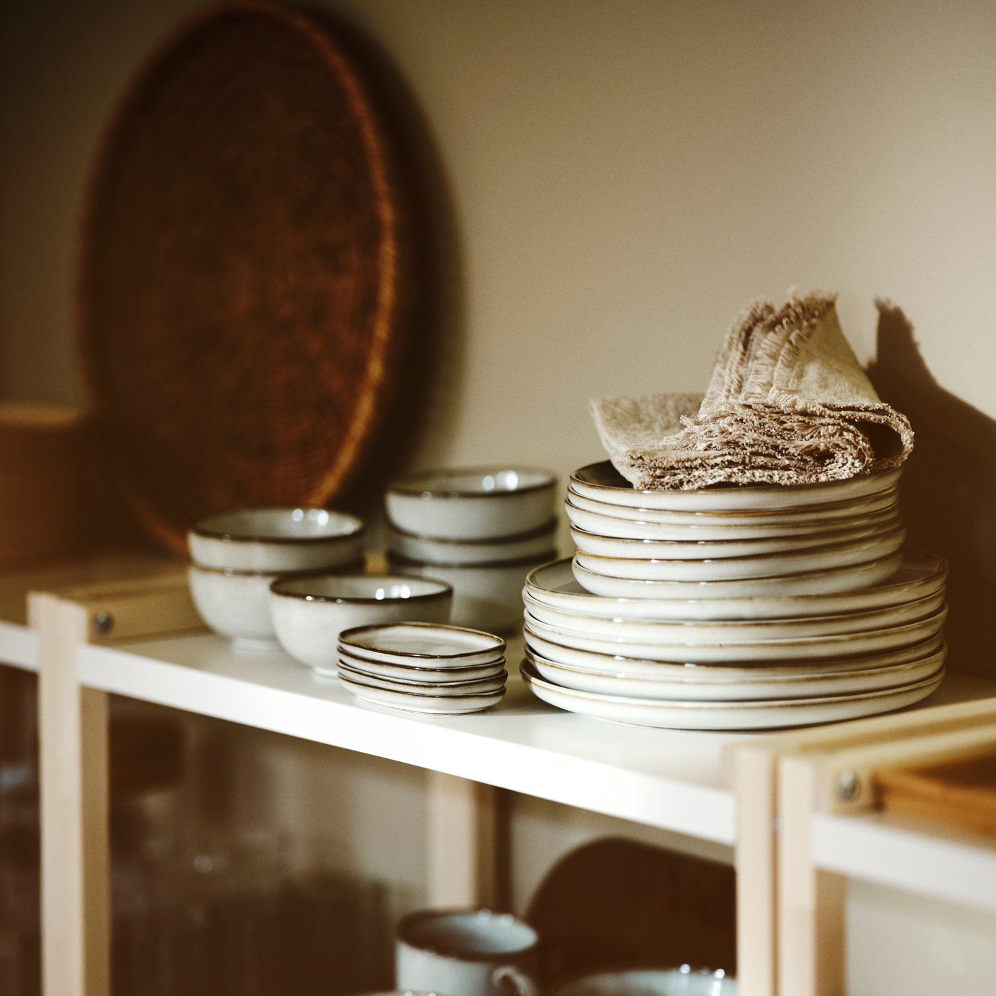 Kuhinja, v kateri so na kup zloženi sivi GLADELIG desertni krožniki, servirne sklede, lončki, namizni prtički, lesena enota za shranjevanje in pladenj.