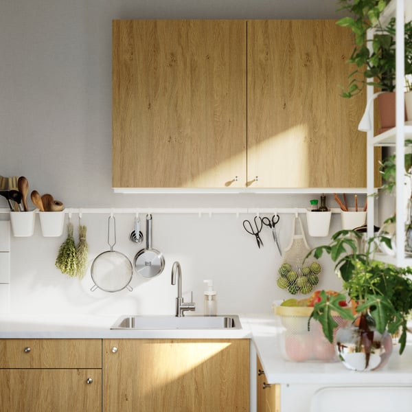 A KNOXHULT wall cabinet with doors in a light wood finish, set in a modern kitchen with utensils and plants.