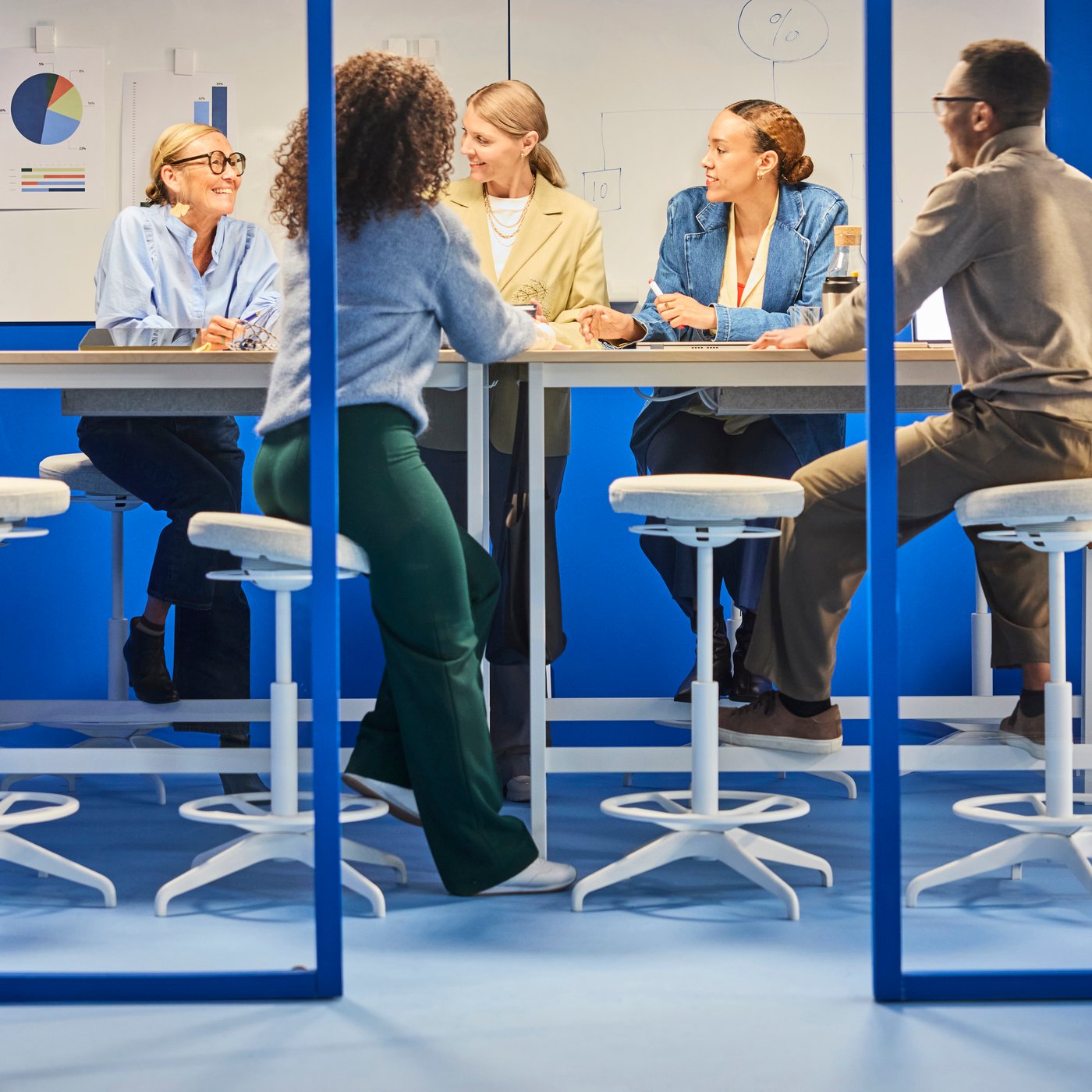 Teammöte runt ett högt bord i ett ljust kontor, människor diskuterar idéer med anteckningar på väggen, sitter på pallar.