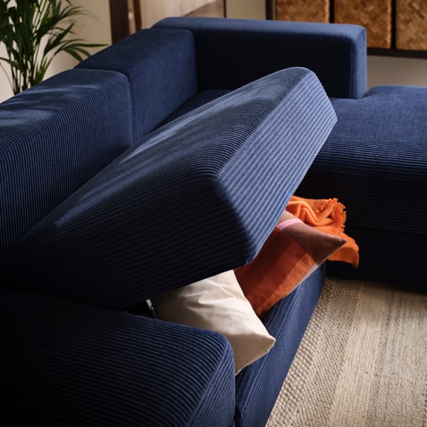 A living room with a dark-blue JÄTTEBO four-seat sofa, the middle seat raised, holding a pair of cushions and a throw within.
