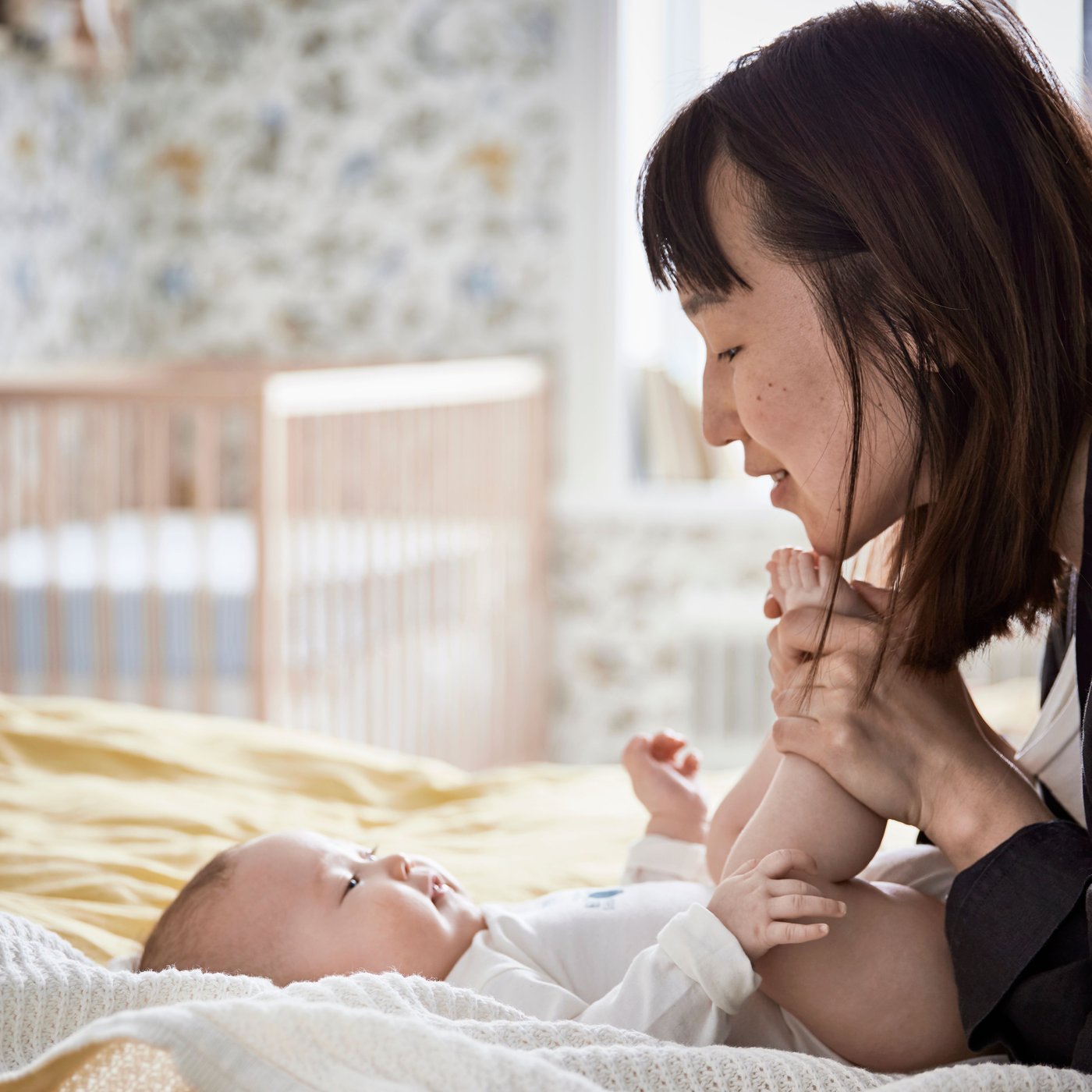 A happy baby lies on a white GULSPARV blanket on top of a bed with her mother who holds her feet and smiles at her.