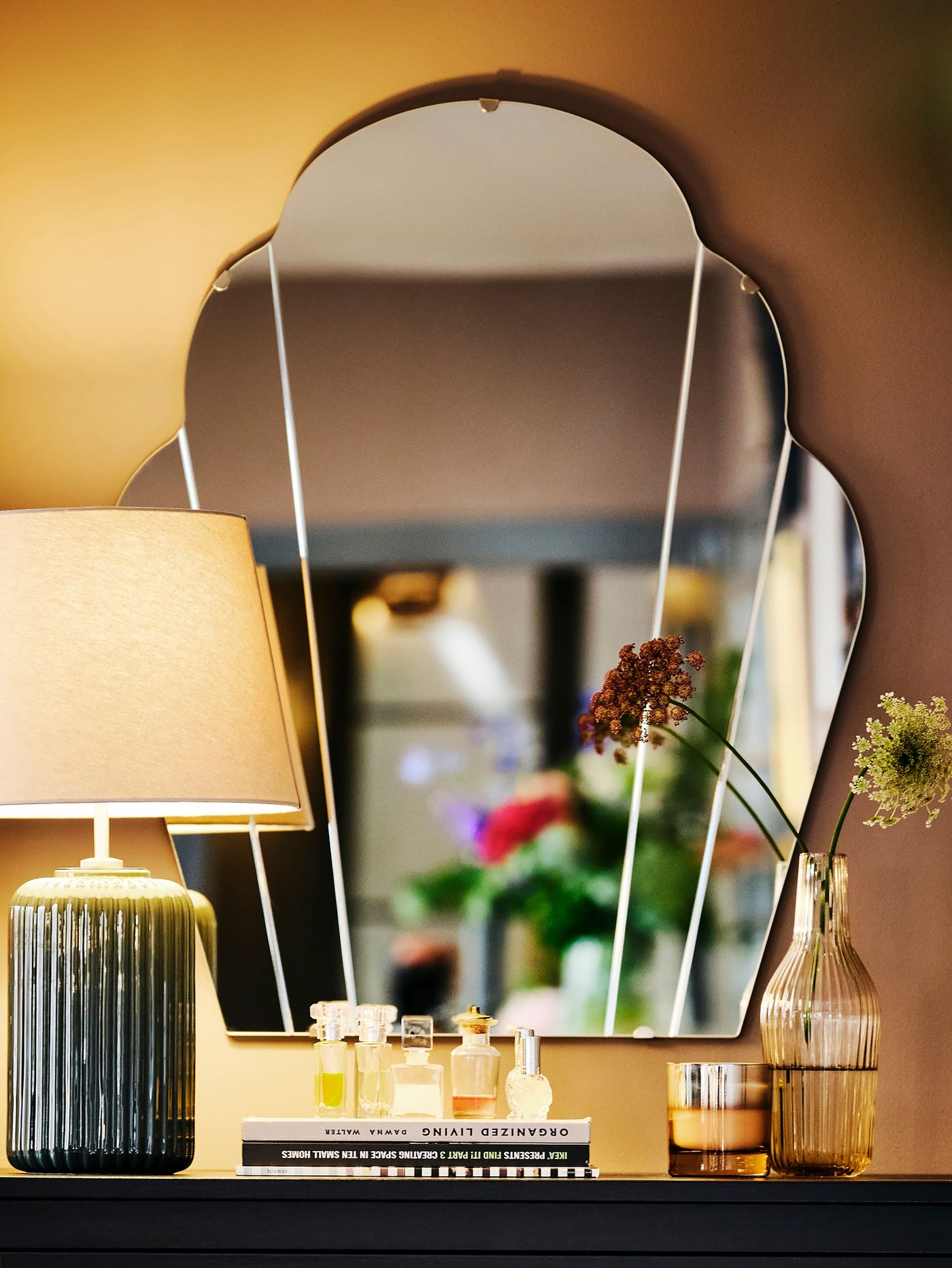 A LOMMARYD mirror is attached to the wall above a cabinet that has a lamp, books, perfume bottles and flowers on top of it.