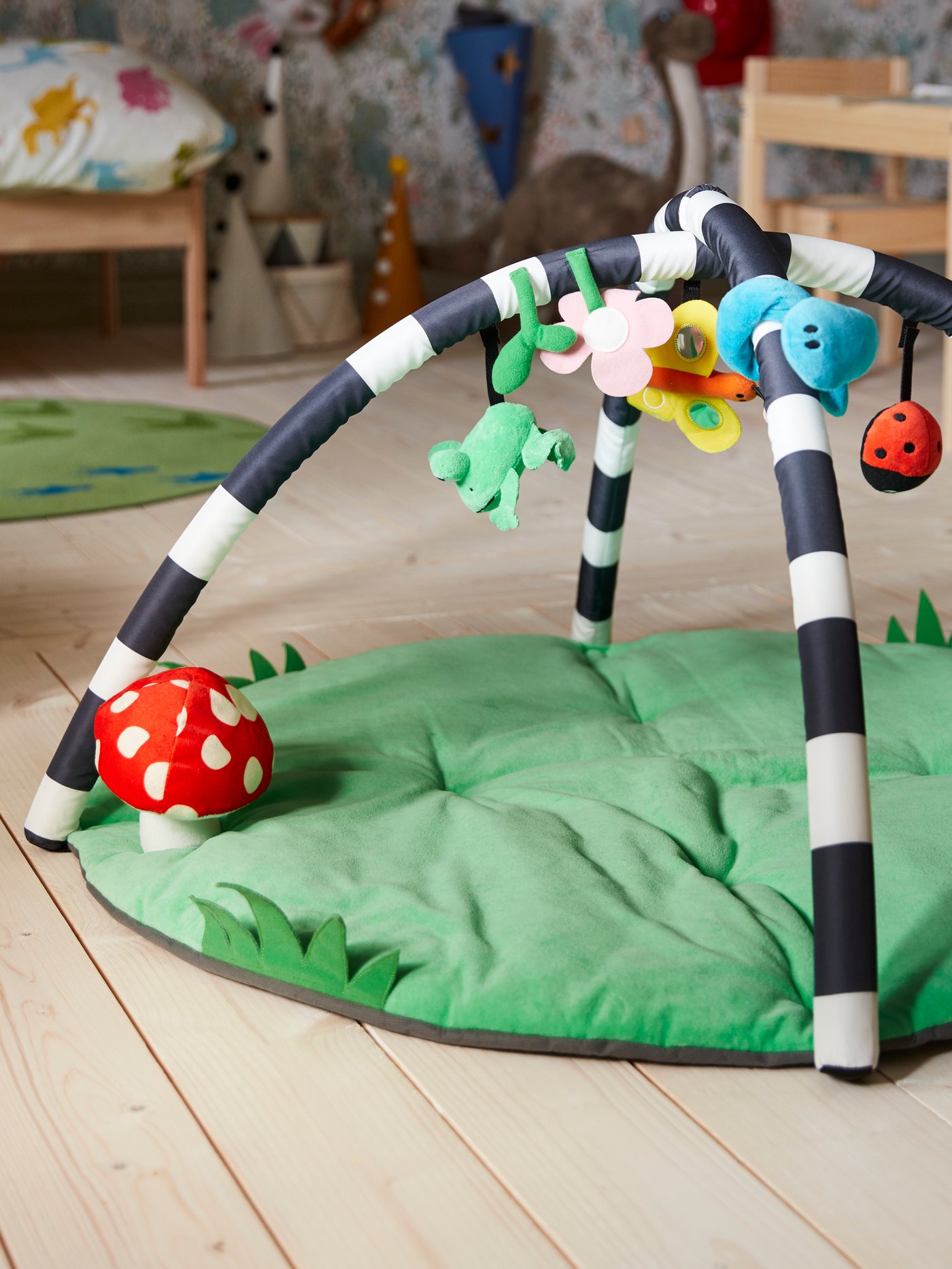 A KLAPPA baby gym on a wooden floor with a green base with a mushroom toy, and ladybug, butterfly and frog toys overhead.