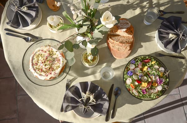 Set dining table with flowers, dessert, salad with edible flowers, bread and water glasses on light tablecloth.