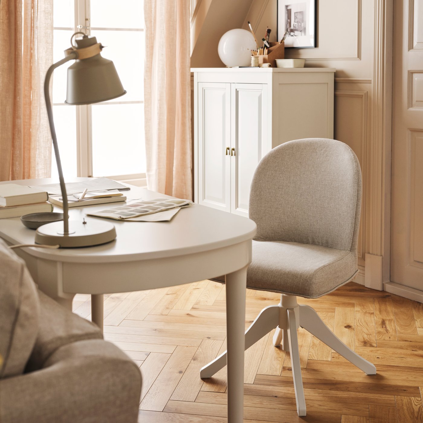 A white ROSENTORP desk and a ROSENTORP swivel chair stand behind a sofa near an off-white FÄRJKARL cabinet in a living room.