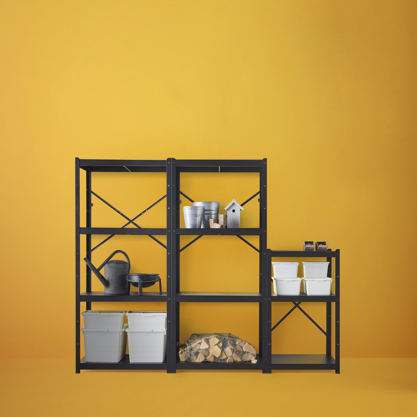 Combinaison d’étagères BROR en noir contre un mur orange clair, avec bûches, boîtes de rangement blanches et outils de jardinage.