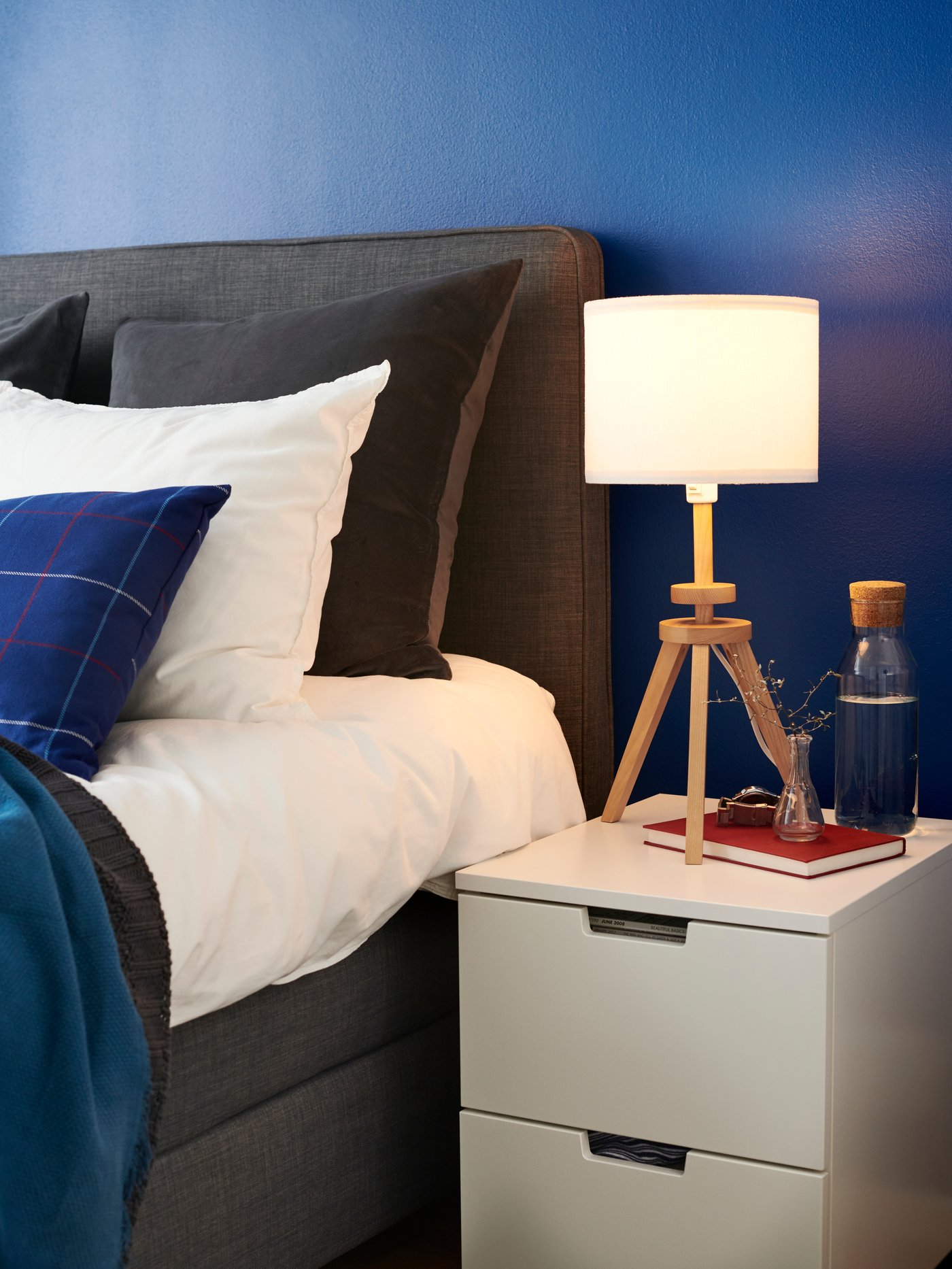 Una cama gris con respaldo NORDLI de tela gris. Al costado de la cama un velador blanco con una lámpara encendida encima